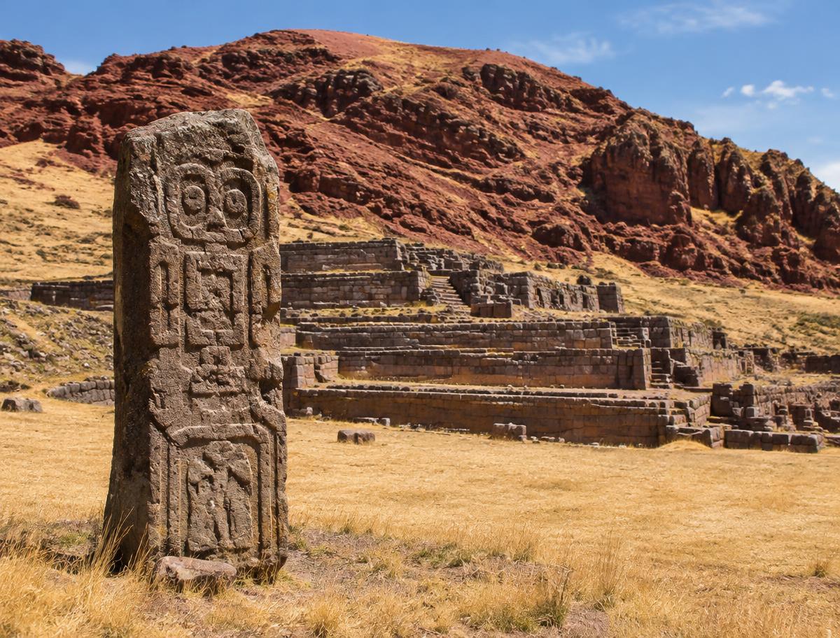 A carved stone stela in front of the terraced ceremonial platforms of Pukará, with a red sandstone hill behind
